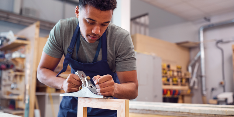 Male Student Studying For Carpentry Apprenticeship At College Using Wood Plane. CREDIT_istock-1218972142.png
