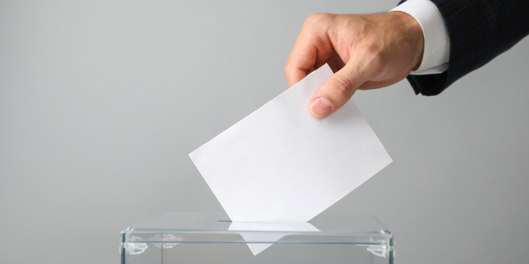 The hand is holding a white envelope or voting slip. iStock-1267315816