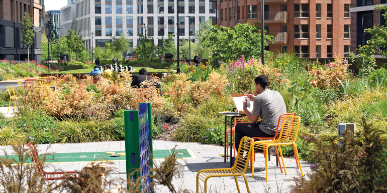 A man checks his phone while enjoying his lunch break in an urban garden adorned with vibrant summer greenery, surrounded by office and apartments.CREDIT_shutterstock_2638583967