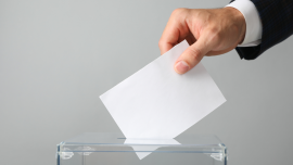 The hand is holding a white envelope or voting slip. iStock-1267315816